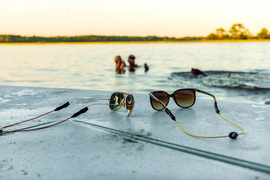 Two pairs of sunglasses with GRAPLRZ sunglass retainers at water's edge and people swimming in the background