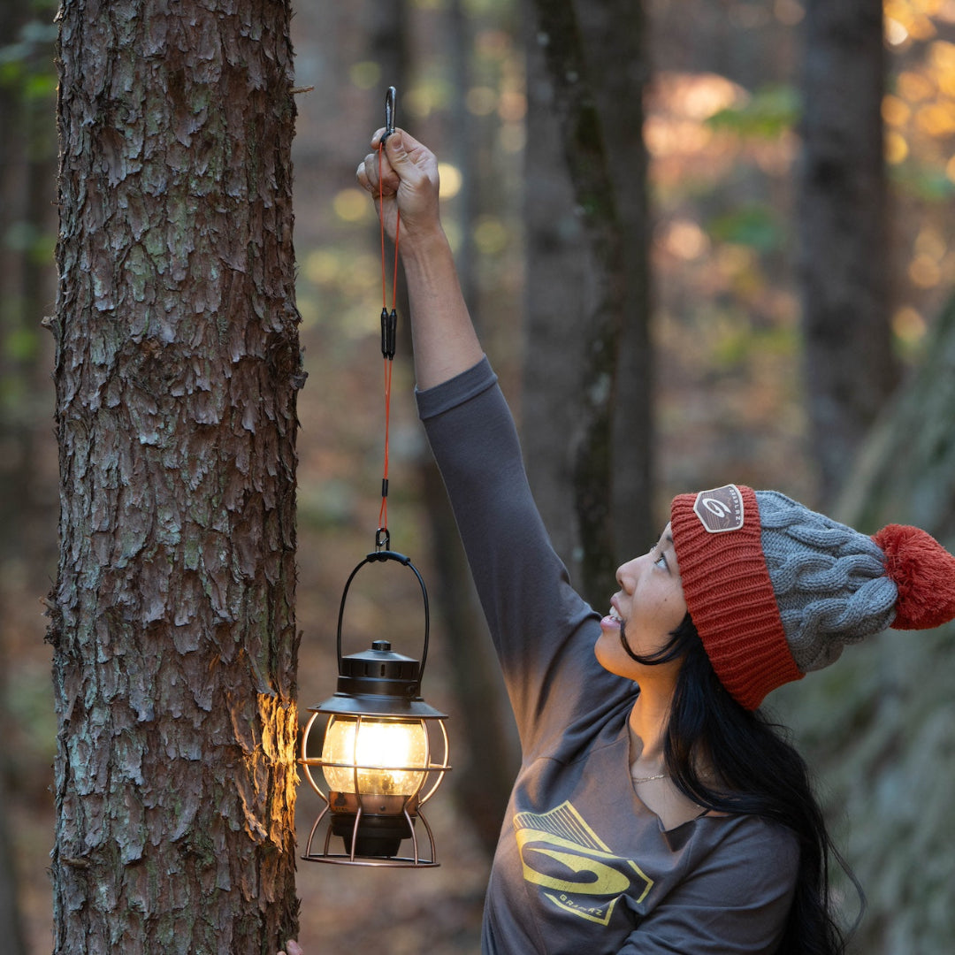 Woman in light grey Chunky Drifter beanie hanging a camping lantern on a tree from orange GRAPLRZ VERZA Multi Use Lanyard