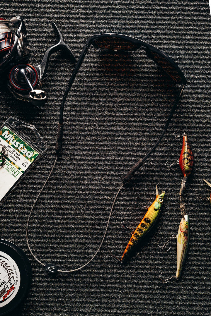 Black sunglasses with dark grey GRAPLRZ sunglass retainers on a black ridged background, surrounded by various fishing items