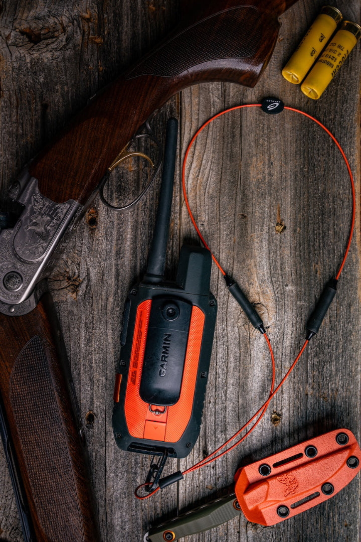 Red GRAPLRZ VERZA Multi Use Lanyard clipped to a walkie talkie on a wood background surrounded by various items