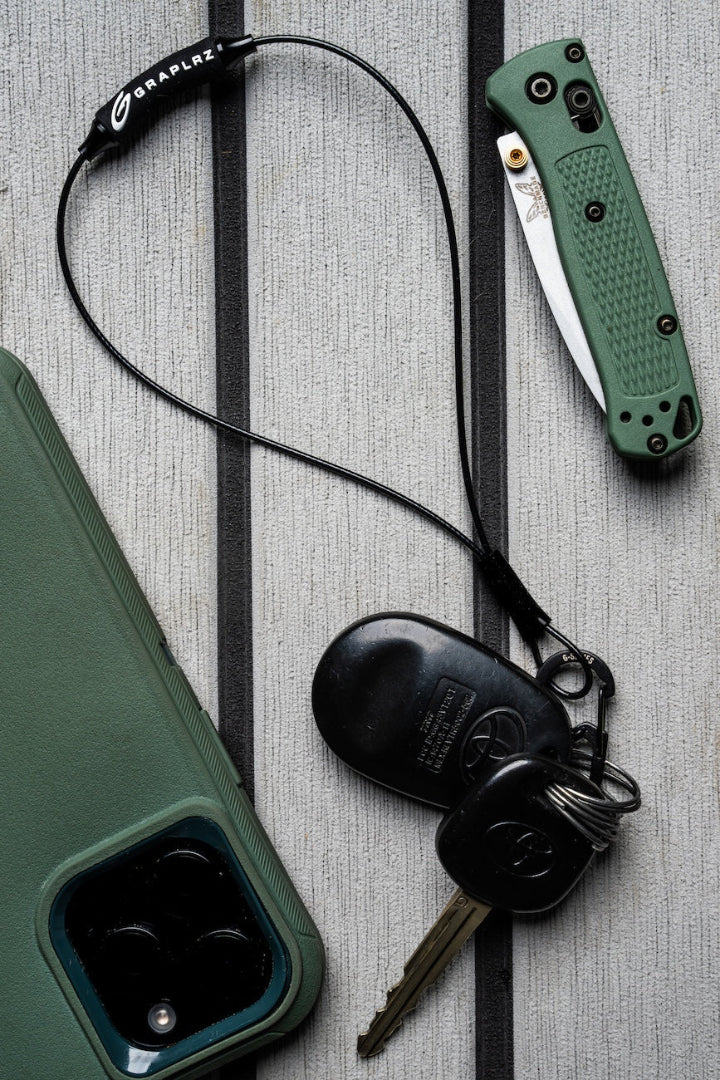 GRAPLRZ Black Verza Mini Utility Lanyard clipped to car keys on wood planks next to a smartphone and pocketknife