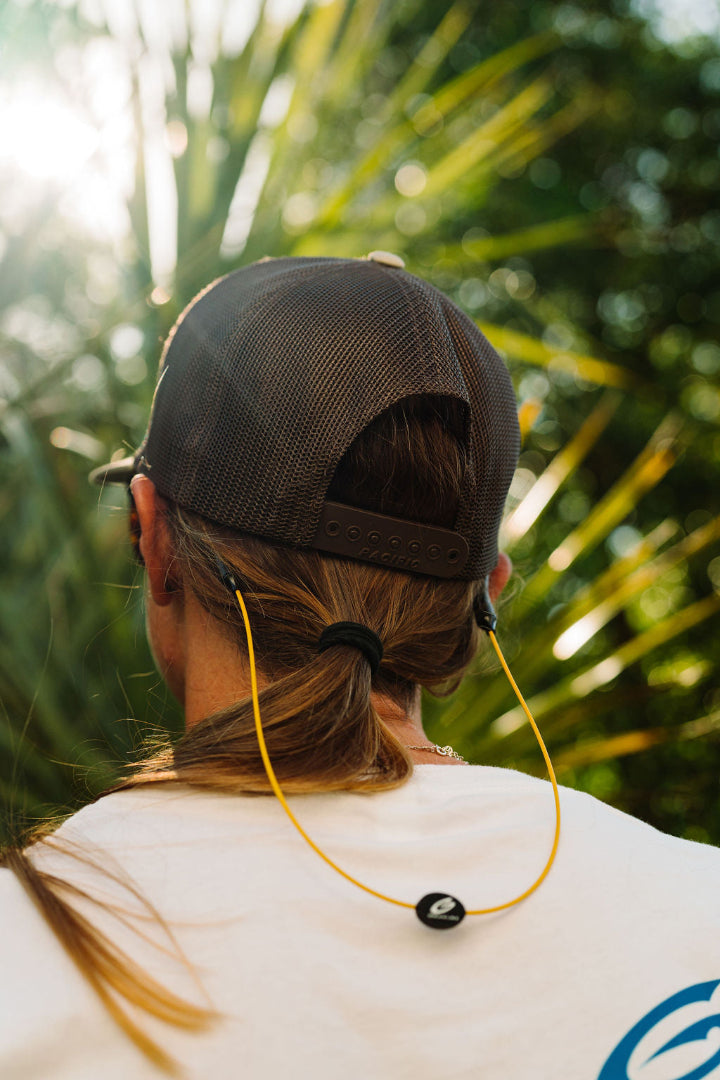 Girl with her back turned wearing a black trucker hat and yellow GRAPLRZ Original Sunglass Retainers in front of tropical leaves