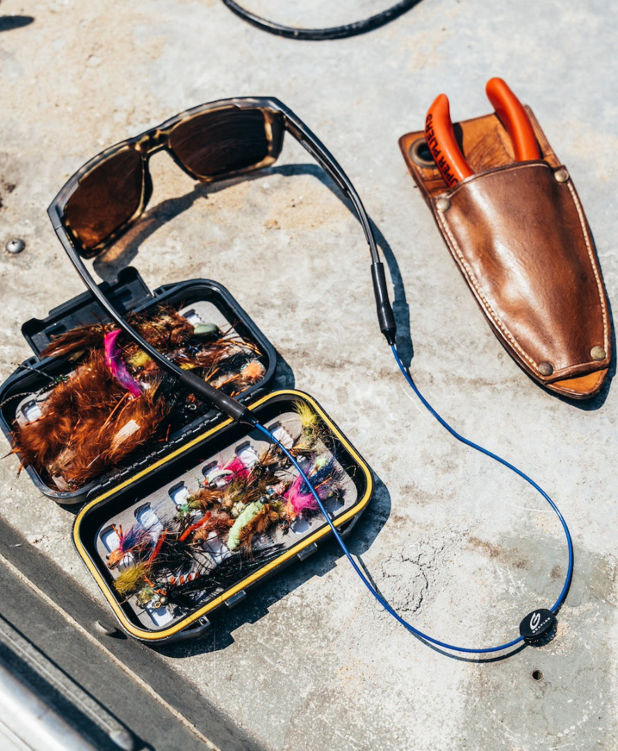Pair of tortoiseshell sunglasses with blue GRAPLRZ sunglass retainers attached, surrounded by various fishing items