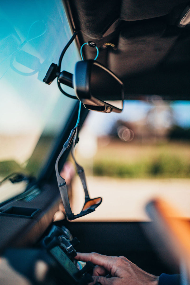 Sunglasses with aqua GRAPLRZ eyeglass retainers hanging from a car rearview mirror